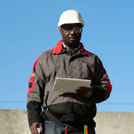 Téléchargez les photos : Travailleur afro-américain en chapeau dur blanc et lunettes de protection se tient sur le chantier de construction, tient dans les mains tablette ordinateur et regarde la caméra - en image libre de droit