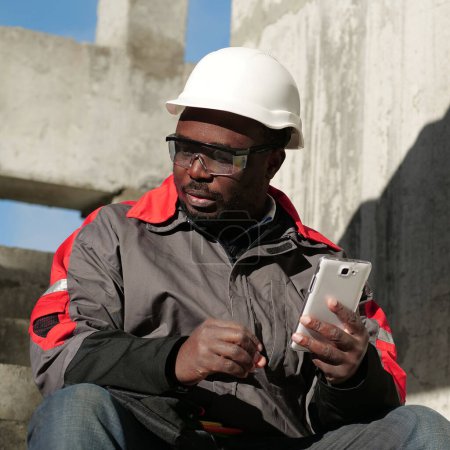 Téléchargez les photos : Travailleur afro-américain en casque dur et lunettes de protection avec smartphone se trouve sur le chantier de construction. Workman tient dans les mains smartphone blanc et pense - en image libre de droit