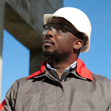 Téléchargez les photos : Travailleur afro-américain en casque et lunettes de protection se tient sur le chantier et regarde autour de lui - en image libre de droit