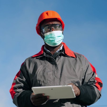 Téléchargez les photos : Travailleur afro-américain dans le masque de protection médicale se trouve sur le chantier de construction avec ordinateur tablette. Workman tient dans les mains une tablette PC blanche et pense - en image libre de droit