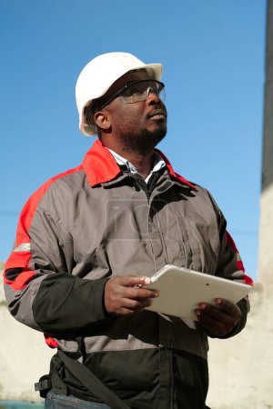 Téléchargez les photos : Travailleur afro-américain en casque dur et lunettes de protection avec tablette se tient sur le chantier et regarde autour - en image libre de droit
