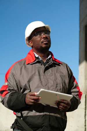 Téléchargez les photos : Travailleur afro-américain en casque dur et lunettes de protection avec tablette se tient sur le chantier de construction - en image libre de droit