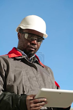 Téléchargez les photos : Ouvrier afro-américain en casque dur et lunettes de protection avec tablette se tient sur le chantier de construction - en image libre de droit