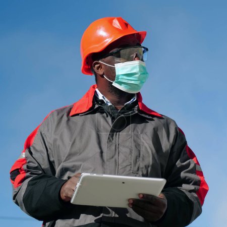 Téléchargez les photos : Travailleur afro-américain dans des stands de masque de protection médicale sur le site de construction avec tablette - en image libre de droit