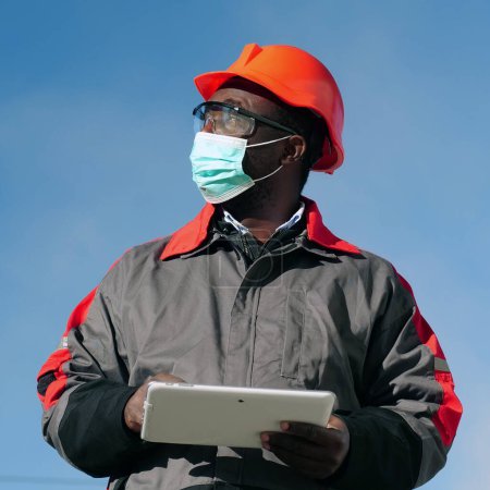 Téléchargez les photos : Travailleur afro-américain en masque de protection médicale se trouve sur le chantier de construction avec ordinateur tablette. Workman tient dans les mains une tablette PC blanche et regarde autour de lui - en image libre de droit