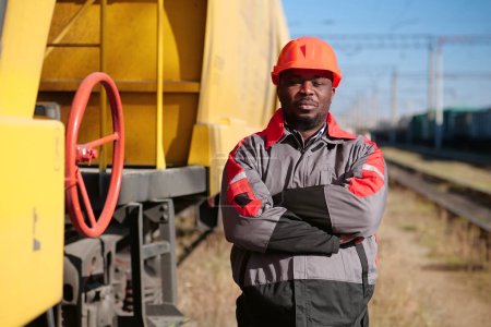 Téléchargez les photos : African american railway worker stands at freight train terminal. Railroad man in uniform and red hard hat look at camera - en image libre de droit