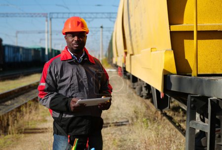Photo for African american railway man with tablet computer at freight train terminal. Railroad man in uniform and red hard hat with computer. Railway employee holds in hands tablet pc and look at the camera - Royalty Free Image
