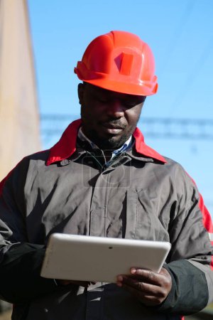 Photo for African american railway man with tablet computer at freight train terminal. Railroad man in uniform and red hard hat with computer. Railway employee holds in hands tablet pc - Royalty Free Image