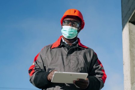 Téléchargez les photos : Travailleur afro-américain dans le masque de protection médicale se trouve sur le chantier de construction avec ordinateur tablette. Workman tient dans les mains une tablette PC blanche et pense - en image libre de droit