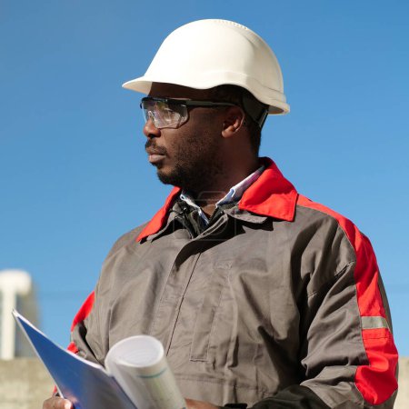 Téléchargez les photos : Un ouvrier afro-américain se tient sur le chantier avec des documents de travail et attend avec impatience - en image libre de droit