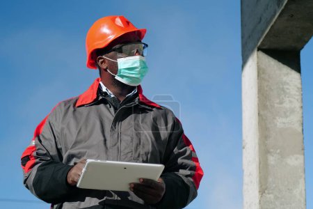 Téléchargez les photos : Travailleur afro-américain dans des stands de masque de protection médicale sur le site de construction avec tablette - en image libre de droit