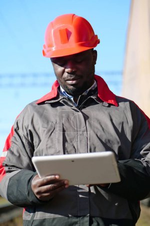 Photo for Railroad man in uniform and red hard hat with computer. Railway employee holds in hands tablet pc. African american railway man with tablet computer at freight train terminal - Royalty Free Image