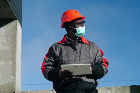 Téléchargez les photos : Travailleur afro-américain en masque protecteur médical se tient sur le chantier de construction et prend des notes dans un ordinateur tablette. Workman tient dans les mains la tablette PC blanche - en image libre de droit