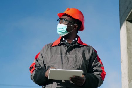 Téléchargez les photos : Travailleur afro-américain en masque de protection médicale se trouve sur le chantier de construction avec ordinateur tablette. Workman tient dans les mains une tablette PC blanche et regarde autour de lui - en image libre de droit