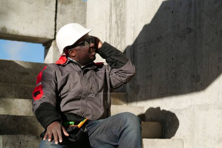 Téléchargez les photos : Un ouvrier afro-américain est assis dans les escaliers du chantier. Il ne se sent pas bien et a mal à la tête, il touche sa tête - en image libre de droit