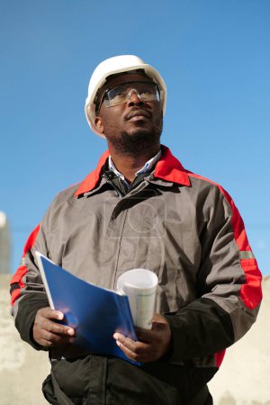 Téléchargez les photos : Un ouvrier afro-américain se tient sur le chantier avec des papiers de travail et regarde autour de lui - en image libre de droit
