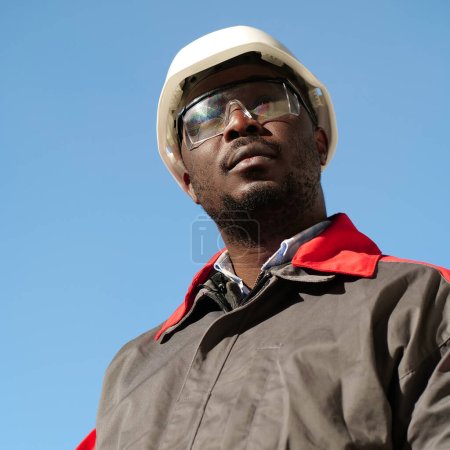 Téléchargez les photos : Travailleur afro-américain en casque et lunettes de protection se tient sur le chantier de construction - en image libre de droit