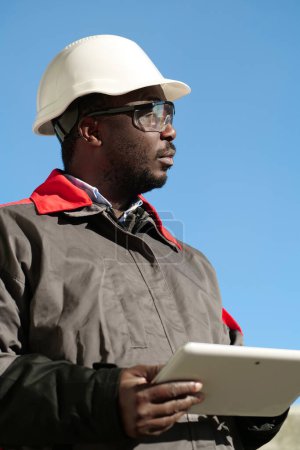 Téléchargez les photos : Travailleur afro-américain en casque dur et lunettes de protection avec tablette se tient sur le chantier et regarde autour - en image libre de droit