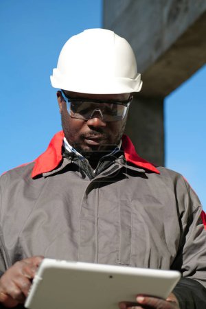 Téléchargez les photos : Travailleur afro-américain en casque dur et lunettes de protection se tient sur le chantier de construction et travaille avec tablette informatique - en image libre de droit