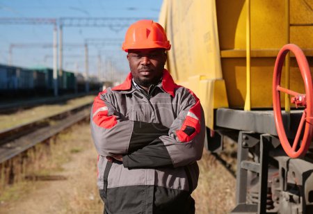 Photo for African american railway man at freight train terminal. Railroad man in uniform and red hard hat look at the camera - Royalty Free Image