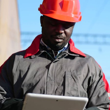 Photo for African american railway man with tablet computer at freight train terminal. Railroad man in uniform and red hard hat with computer. Railway employee holds in hands tablet pc - Royalty Free Image