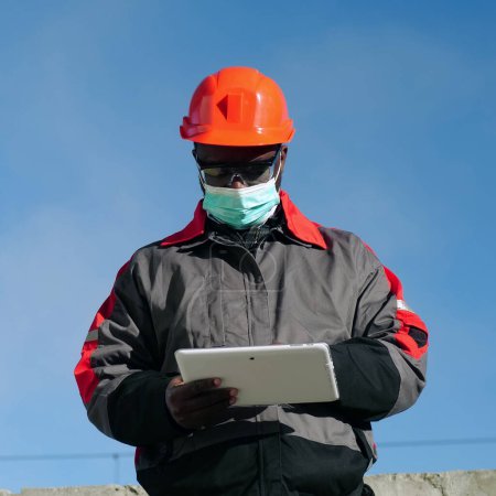 Téléchargez les photos : Travailleur afro-américain en masque protecteur médical se tient sur le chantier de construction et prend des notes dans un ordinateur tablette. Workman tient dans les mains la tablette PC blanche - en image libre de droit