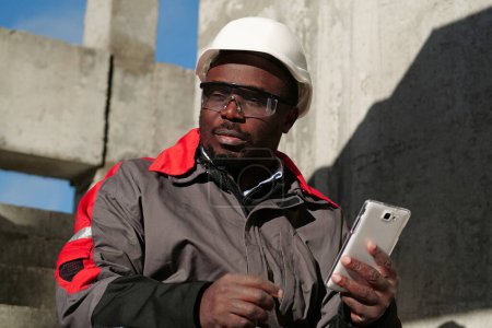 Téléchargez les photos : Travailleur afro-américain en casque dur et lunettes de protection avec smartphone se trouve sur le chantier de construction. Workman tient dans les mains smartphone blanc - en image libre de droit