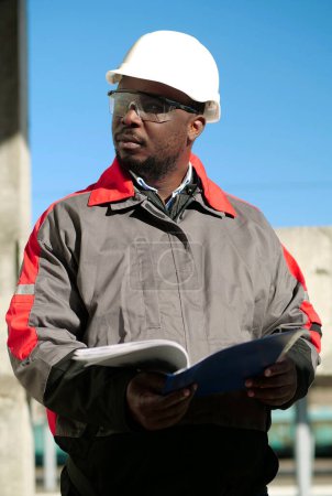 Téléchargez les photos : Un ouvrier afro-américain se tient sur le chantier avec des papiers de travail et regarde autour de lui - en image libre de droit