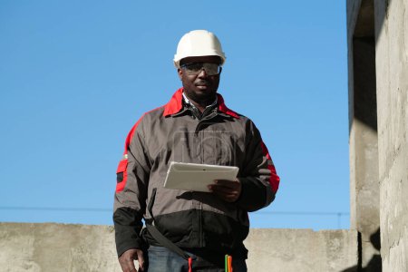 Téléchargez les photos : Travailleur afro-américain en chapeau dur blanc et lunettes de protection se tient sur le chantier de construction, tient dans les mains tablette ordinateur et regarde la caméra - en image libre de droit