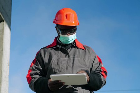 Téléchargez les photos : Travailleur afro-américain en masque de protection médicale se tient sur le chantier de construction et regarde dans l'ordinateur tablette - en image libre de droit