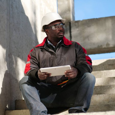 Téléchargez les photos : Travailleur afro-américain fatigué avec les yeux fermés en casque blanc assis sur le chantier de construction et tient dans les mains tablette PC - en image libre de droit