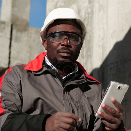 Téléchargez les photos : Travailleur afro-américain en casque dur et lunettes de protection avec smartphone se trouve sur le chantier de construction. Workman tient dans les mains smartphone blanc et pense - en image libre de droit