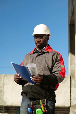 Téléchargez les photos : Un ouvrier afro-américain se tient sur le chantier et regarde les papiers de travail - en image libre de droit
