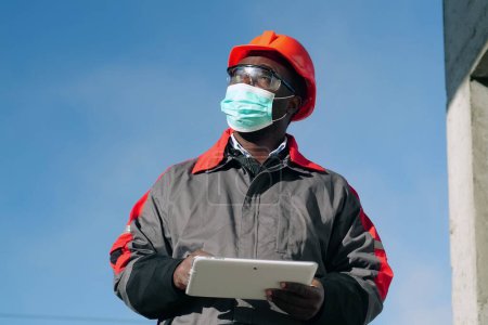 Téléchargez les photos : Travailleur afro-américain dans le masque de protection médicale se tient à la construction avec tablette informatique. Workman tient dans les mains une tablette PC blanche et regarde autour de lui - en image libre de droit