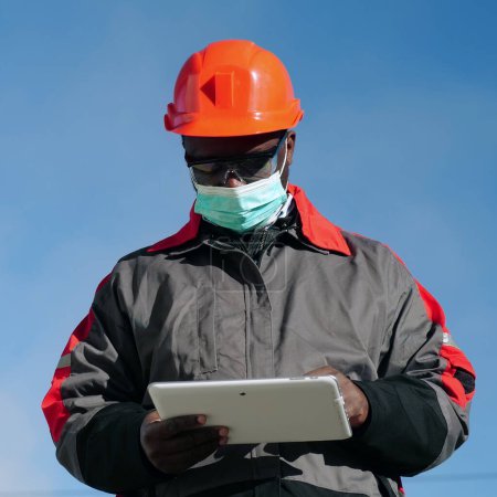 Téléchargez les photos : Un travailleur afro-américain portant un masque protecteur médical se tient sur le chantier et prend des notes sur sa tablette. Workman tient dans les mains la tablette PC blanche - en image libre de droit