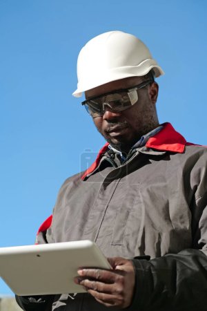 Téléchargez les photos : Travailleur afro-américain en casque dur et lunettes de protection avec tablette se tient sur le chantier de construction - en image libre de droit
