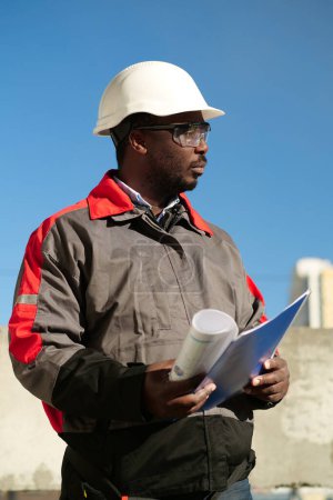 Téléchargez les photos : Un ouvrier afro-américain se tient sur le chantier avec des documents de travail et attend avec impatience - en image libre de droit