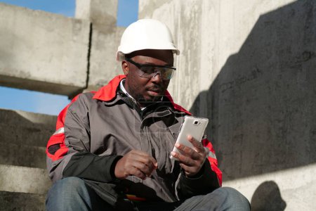 Téléchargez les photos : Travailleur afro-américain en casque dur et lunettes de protection avec smartphone se trouve sur le chantier de construction. Workman regarde dans son smartphone - en image libre de droit
