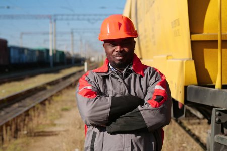 Photo for African american railway man at freight train terminal. Railroad man in uniform and red hard hat look at the camera - Royalty Free Image