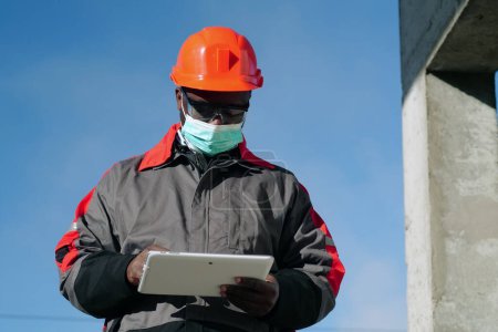 Téléchargez les photos : Un travailleur afro-américain portant un masque protecteur médical se tient sur le chantier et prend des notes sur sa tablette. Workman tient dans les mains la tablette PC blanche - en image libre de droit