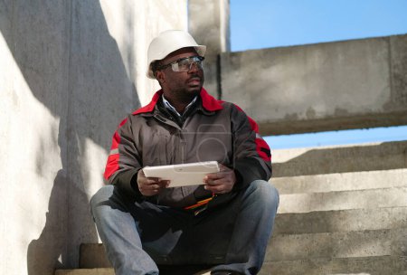 Téléchargez les photos : Ouvrier afro-américain fatigué, les yeux à moitié fermés, en casque blanc, assis sur le chantier et tenant dans les mains une tablette PC - en image libre de droit
