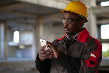 Téléchargez les photos : Un ouvrier afro-américain en casque jaune avec smartphone se tient sur le chantier. Travailleur tient dans les mains smartphone - en image libre de droit