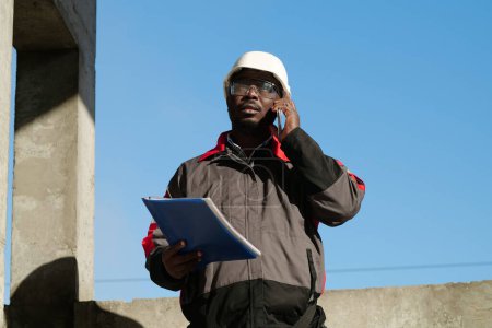 Téléchargez les photos : Travailleur afro-américain avec des documents de travail parle sur smartphone. Gestionnaire de travaux afro-américain occupé en casque dur avec des documents de travail se tient sur le chantier de construction et parle sur smartphone - en image libre de droit