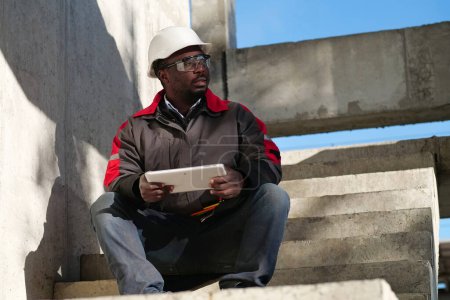 Téléchargez les photos : Travailleur afro-américain fatigué avec les yeux à moitié fermés en casque blanc assis sur le chantier de construction et tient dans les mains tablette PC - en image libre de droit