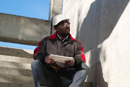 Téléchargez les photos : Travailleur afro-américain en chapeau dur blanc se trouve sur le site de construction et tient dans les mains tablette PC - en image libre de droit