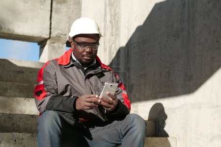 Téléchargez les photos : Travailleur afro-américain en casque dur et lunettes de protection avec smartphone se trouve sur le chantier de construction. Workman tient dans les mains smartphone blanc et regarde la caméra - en image libre de droit
