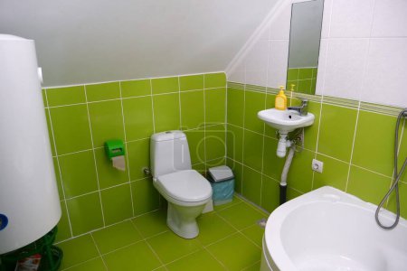 Téléchargez les photos : Intérieur de la salle de toilettes avec cuvette de toilette blanche avec couvercle fermé, chaudière électrique pour le chauffage de l'eau, douche, lavabo et carrelage mural et plancher vert - en image libre de droit