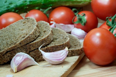 A cutting board displaying slices of bread beside a pile of garlic and tomatoes.