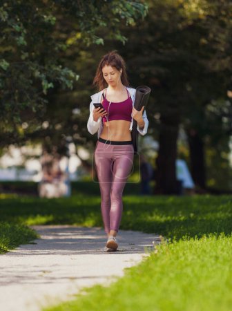 Photo for Slim fit caucasian woman in colorful fitness outfit walking in park with yoga mat and smartphone - Royalty Free Image