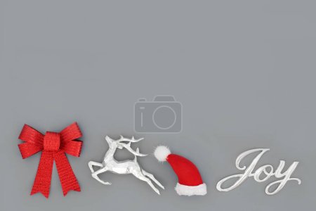 Foto de Concepto de alegría navideña con signo, sombrero de santa, reno y lazo rojo sobre fondo gris. Decoraciones y símbolos festivos de árboles. Diseño minimalista abstracto para vacaciones de Navidad y Año Nuevo. - Imagen libre de derechos